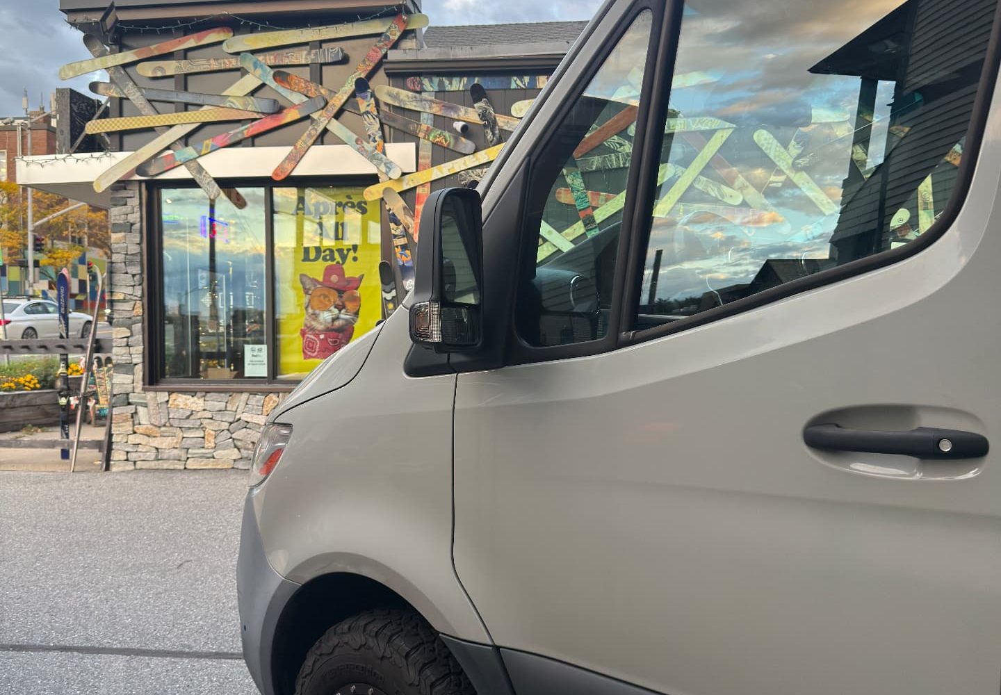 Sprinter front wheel and cabin outside a ski shop with crossed vintage skis and an Après All Day poster